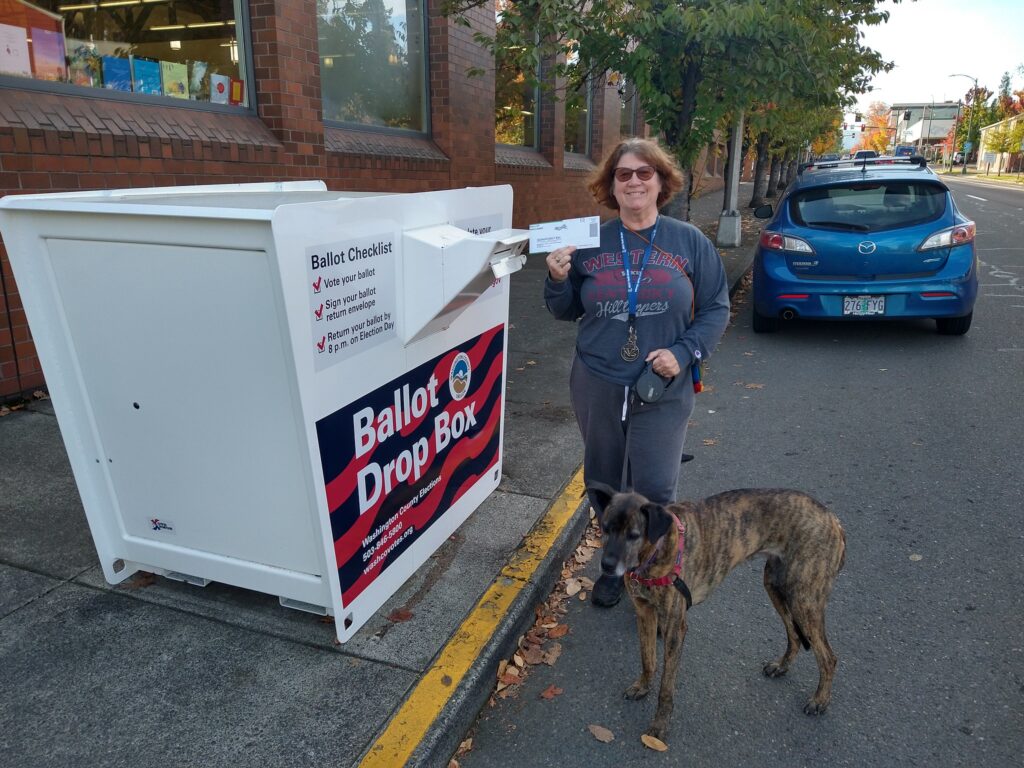 A woman holds her ballot up proudly and is standing in front of the ballot drop box where she will drop it. She has a dog on a leash next to her. 